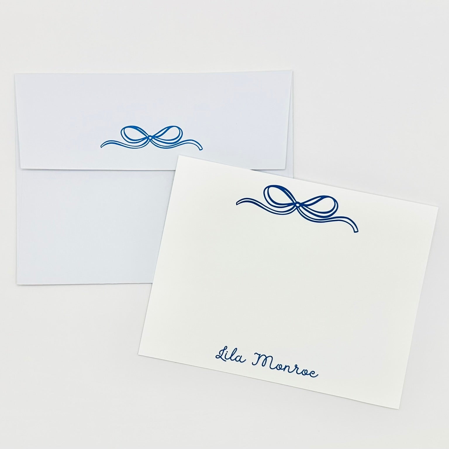 White stationery flat card with blue decorative bow and personalized name 'Lila Monroe' text, paired with a matching envelope on a white background.