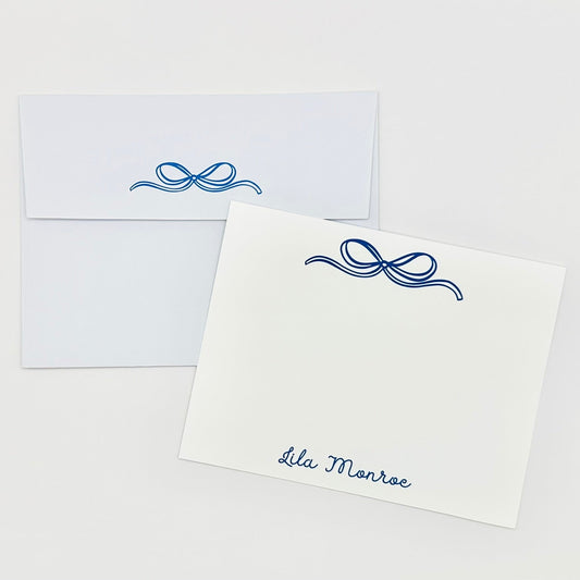 White stationery flat card with blue decorative bow and personalized name 'Lila Monroe' text, paired with a matching envelope on a white background.